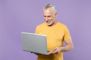 Smiling elderly gray-haired mustache bearded man in casual yellow t-shirt isolated on violet background studio portrait. People lifestyle concept. Mock up copy space. Working on laptop pc computer.