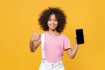 Smiling african american kid girl 12-13 years old in pink t-shirt isolated on yellow background. Childhood concept. Mock up copy space. Hold mobile phone with blank empty screen, showing thumb up.