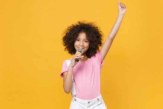 Funny little african american kid girl 12-13 years old in pink t-shirt isolated on yellow wall background studio. Childhood lifestyle concept. Mock up copy space. Sing song in microphone rising hand.