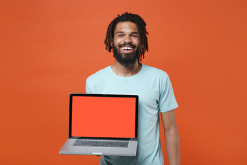 Smiling young african american man guy wearing blue casual t-shirt posing isolated on orange background. People lifestyle concept. Mock up copy space. Hold laptop pc computer with blank empty screen.