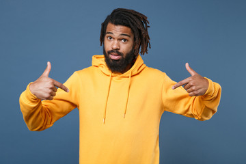 Amazed young african american man guy wearing yellow streetwear hoodie posing isolated on blue wall background studio portrait. People emotions lifestyle concept. Pointing index fingers on himself.
