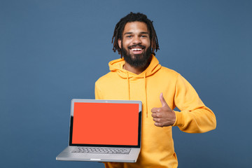 Smiling young african american man guy in yellow hoodie isolated on blue background. People lifestyle concept. Mock up copy space. Hold laptop pc computer with blank empty screen, showing thumb up.