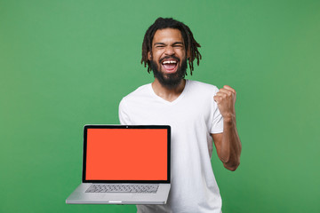 Happy young african american man 20s in white t-shirt posing hold laptop pc computer with blank empty screen with mock up copy space doing winner gesture isolated on green background studio portrait.