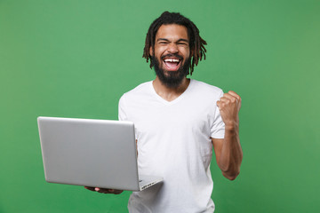 Joyful young african american man guy with dreadlocks 20s wearing white casual t-shirt posing hold laptop pc computer doing winner gesture isolated on green color wall background studio portrait.