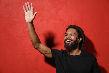Cheerful joyful young african american man guy with dreadlocks 20s in black casual t-shirt posing waving greeting with hand as notices someone isolated on bright red color background studio portrait.