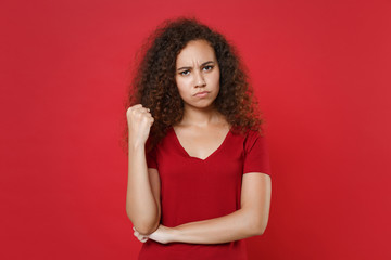 Displeased young african american woman girl in casual t-shirt posing isolated on red wall background studio portrait. People sincere emotions lifestyle concept. Mock up copy space. Clenching fist.