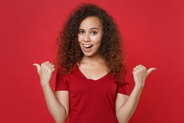 Amazed young african american woman girl in casual t-shirt posing isolated on red wall background studio portrait. People sincere emotions lifestyle concept. Mock up copy space. Pointing thumbs aside.