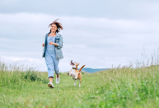 Happy smiling jogging female with fluttering hairs and her beagle dog running and looking at eyes. Walking by meadow grass path in nature with pets, healthy active people lifestyle concept image.