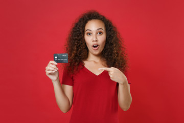 Amazed young african american woman girl in casual t-shirt posing isolated on red background studio portrait. People lifestyle concept. Mock up copy space. Pointing index finger on credit bank card.