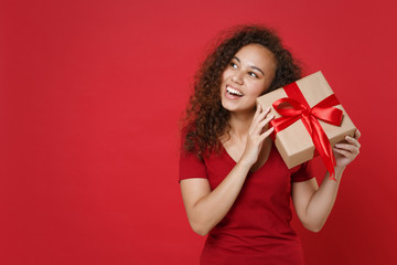 Curious funny young african american woman girl in casual t-shirt posing isolated on red background. St. Valentine's Day, Women's Day, birthday, holiday concept. Hold present box with gift ribbon bow.