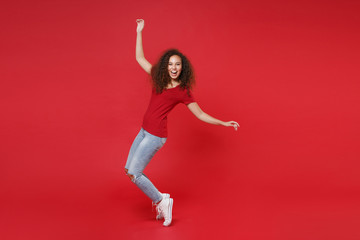 Fototapeta premium Full length portrait of laughing young african american girl in casual t-shirt isolated on red background studio. People lifestyle concept. Mock up copy space. Dancing, standing on toes, rising hands.
