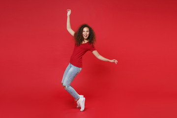 Full length portrait of laughing young african american girl in casual t-shirt isolated on red background studio. People lifestyle concept. Mock up copy space. Dancing, standing on toes, rising hands.