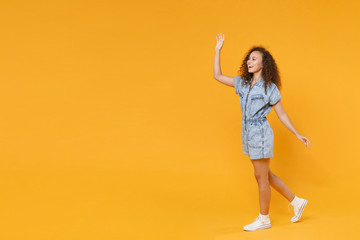 Fototapeta na wymiar Full length portrait side view of funny young african american girl in denim clothes isolated on yellow background studio. People lifestyle concept. Waving and greeting with hand as notices someone.