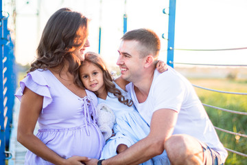 Fototapeta premium happy young family are hugging on the bridge in the sunset light. husband and a little girl touching pregnant wife's belly