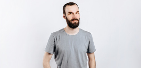 Obraz premium A handsome brutal brunette man with a beard in a gray shirt smiles and looks at the camera. Place for advertising on a gray background.