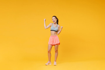 Full length portrait happy young fitness woman in sportswear working out isolated on yellow background in studio. Workout sport motivation lifestyle concept. Mock up copy space. Doing winner gesture.