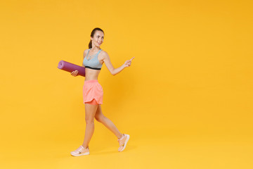 Fototapeta na wymiar Full length portrait of smiling young fitness woman girl in sportswear working out isolated on yellow background. Workout sport motivation lifestyle concept. Hold yoga mat pointing index finger aside.