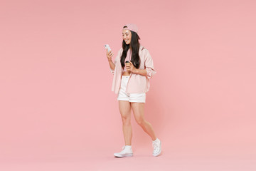 Fototapeta na wymiar Full length portrait of pretty asian girl in casual clothes, cap isolated on pink background. People lifestyle concept. Mock up copy space. Using mobile cell phone, hold paper cup of coffee or tea.