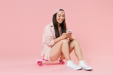 Full length portrait of smiling young asian girl in casual clothes, cap isolated on pastel pink background . People lifestyle concept. Sit on skateboard, using mobile cell phone, typing sms message.