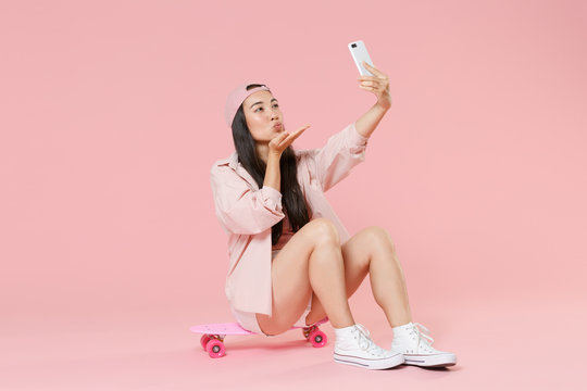 Full length portrait of cute young asian woman girl in cap isolated on pastel pink background. People lifestyle concept. Sit on skateboard, doing selfie shot on mobile phone, blowing sending air kiss.
