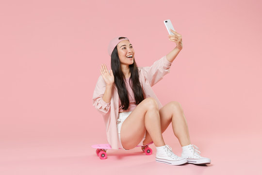 Full length portrait of funny young asian woman girl in cap isolated on pastel pink wall background. People lifestyle concept. Sit on skateboard, doing selfie shot on mobile phone, greeting with hand.