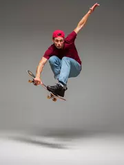 Schilderijen op glas Cool young guy skateboarder jumps on skateboard in studio on gray background. Photography about skateboarding tricks © Georgii