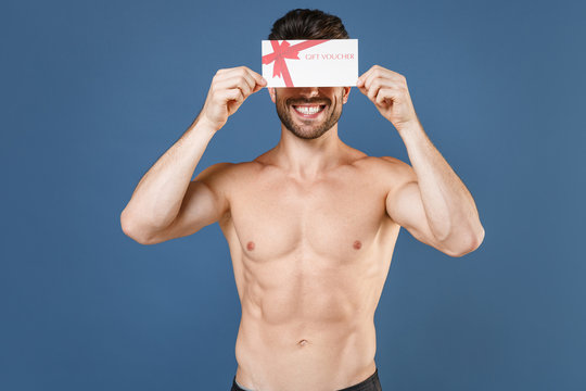 Smiling young bearded fitness sporty strong guy bare-chested muscular sportsman isolated on blue wall background studio. Workout sport motivation lifestyle concept. Covering eye with gift certificate.