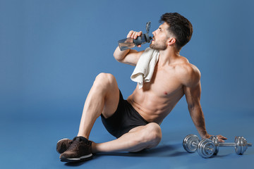 Full length portrait young fitness sporty strong guy bare-chested sportsman isolated on blue background. Workout sport motivation lifestyle concept. Sit on floor with towel on shoulder drinking water.