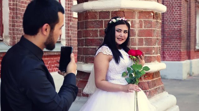 A man takes a photo of a girl with roses on his phone. Wedding photo session. Walk of the bride and groom. A young couple at a red brick column. 4K.