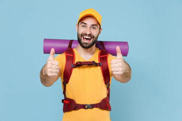 Cheerful traveler young man in yellow casual t-shirt, cap with backpack isolated on blue background studio. Tourist traveling on weekend getaway. Tourism discovering hiking concept. Showing thumbs up.
