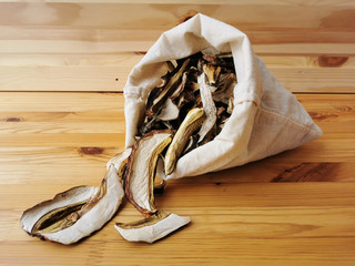 Close-up of dried porcini mushrooms and boletus mushrooms falling out of a white canvas bag on a wooden table against a background of a wooden wall.