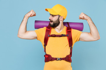 Strong traveler young man in yellow casual t-shirt, cap with backpack isolated on blue background. Tourist traveling on weekend getaway. Tourism discovering hiking concept. Showing biceps, muscles.