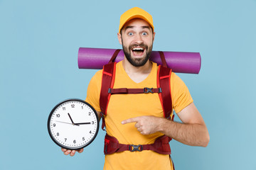 Excited young traveler man in yellow casual t-shirt cap backpack isolated on blue background. Tourist traveling on weekend getaway. Tourism discovering hiking concept. Pointing index finger on clock.