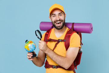 Funny young traveler man in yellow casual t-shirt cap backpack isolated on blue background. Tourist traveling on weekend getaway. Tourism discovering hiking concept. Hold world globe magnifying glass.