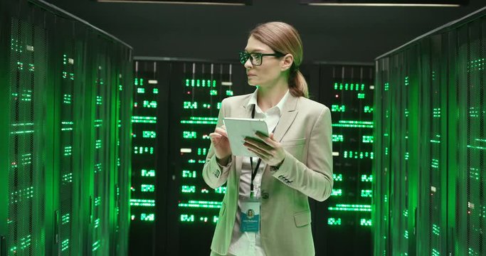 Caucasian beautiful woman in glasses walking at servers with tablet device, tapping and checking big data processors Female analytic in data storage working on digital information security of database