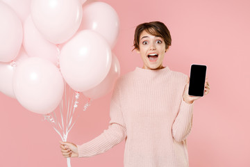 Surprised young woman girl in casual sweater isolated on pastel pink background. Birthday holiday party concept. Mock up copy space. Celebrating hold air balloons mobile phone with blank empty screen.