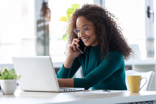 Beautiful young business woman talking on mobile phone while working with her laptop in the office.