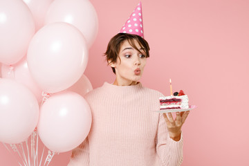 Young brunette woman girl in casual sweater birthday hat isolated on pastel pink background. Birthday holiday party people emotions concept. Celebrating hold air balloons blowing out candle on cake.