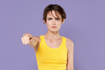 Strict young brunette woman girl in yellow casual tank top posing isolated on pastel violet wall background studio portrait. People sincere emotions lifestyle concept. Pointing index finger on camera.