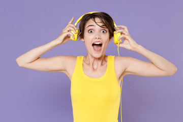Shocked young brunette woman girl wearing yellow casual tank top posing isolated on pastel violet background studio portrait. People sincere emotions lifestyle concept. Listen music with headphones.