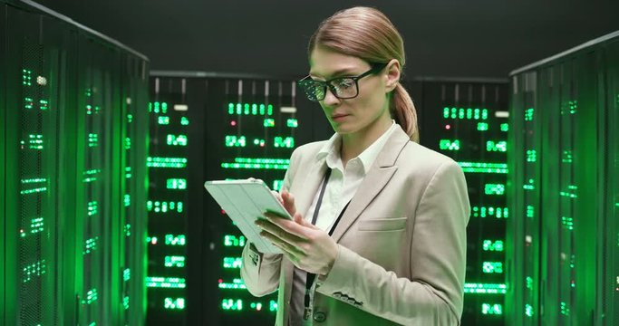 Caucasian young beautiful woman in glasses walking at servers with tablet device, tapping and checking big data processors. Female analytic in data storage working on digital security of database.