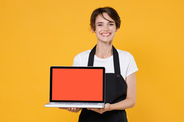 Smiling young female woman 20s barista bartender barman employee in t-shirt apron hold laptop pc computer with blank empty screen mock up copy space isolated on yellow wall background studio portrait.