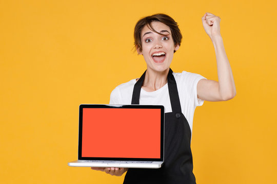 Excited young woman 20s barista bartender barman employee in apron hold laptop computer with blank empty screen mock up copy space doing winner gesture isolated on yellow background studio portrait.