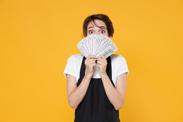 Shocked young female woman 20s barista bartender barman employee in t-shirt apron posing covering face with fan of cash money in dollar banknotes isolated on yellow wall background studio portrait.
