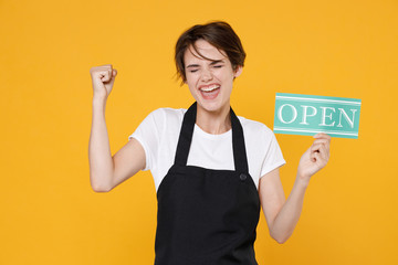 Joyful young female woman 20s barista bartender barman employee in t-shirt apron hold sign with OPEN title clenching fist doing winner gesture isolated on yellow color wall background studio portrait.