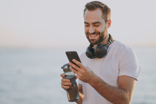 Portrait of smiling young bearded fitness athletic man guy 20s in white t-shirt with headphones posing training resting using mobile cell phone hold bottle of water at sunrise over the sea outdoors.