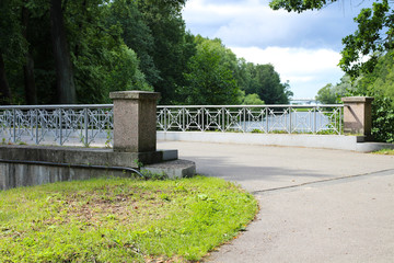 The road to the gray granite bridge over the river with forged metal railings against the sky with clouds.