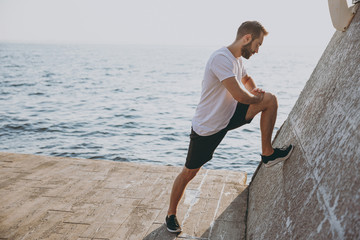 Full length portrait side view of attractive young bearded athletic man guy 20s in white t-shirt posing training doing stretching exercising for legs warming up at sunrise over the sea outdoors.