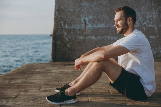 Full length portrait side view of smiling handsome young athletic man guy 20s in casual white t-shirt black shorts posing resting after training looking aside at sunrise over the sea outdoors.