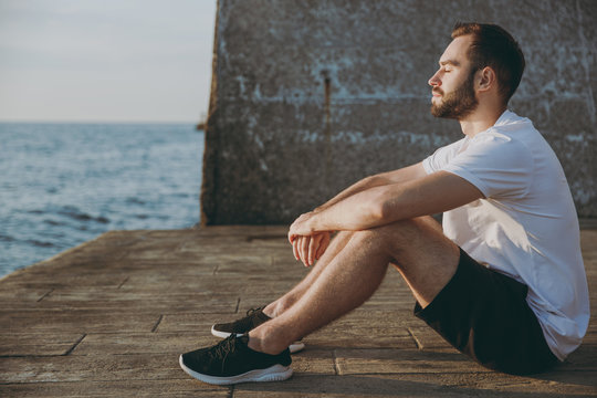 Full length portrait side view of relaxed handsome young athletic man guy 20s in casual white t-shirt black shorts posing resting after training keeping eyes closed at sunrise over the sea outdoors.
